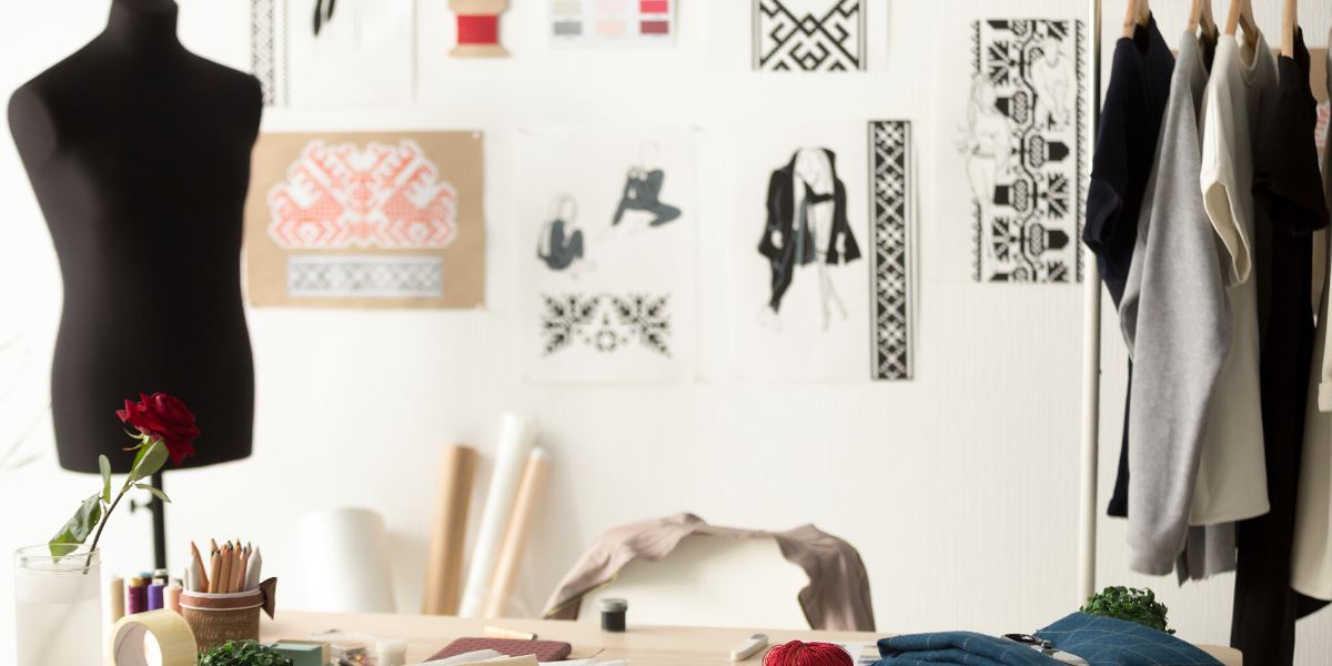 A fashion designer's workspace. Framed by dress form and clothing rack with tops and bottoms. In between, a desk with spools of thread, colored pencils and fabric swatches. Behind, a wall with posters of geometric designs and garment illustrations.