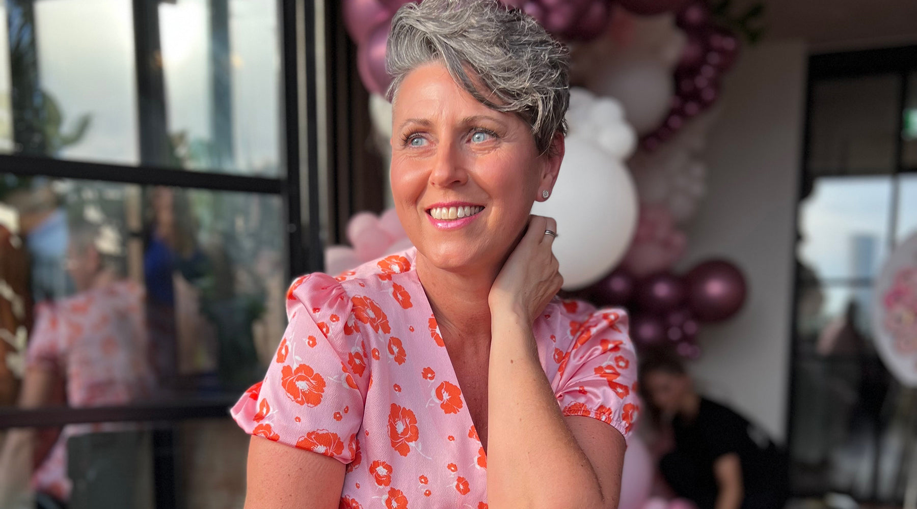 Sandie Roberts is wearing a warm pink flowy summer dress with an orange floral pattern on it. Her left hand is holding the back of her neck and she has a nice smile. Behind her are pink and purple balloons. She looks beautiful.