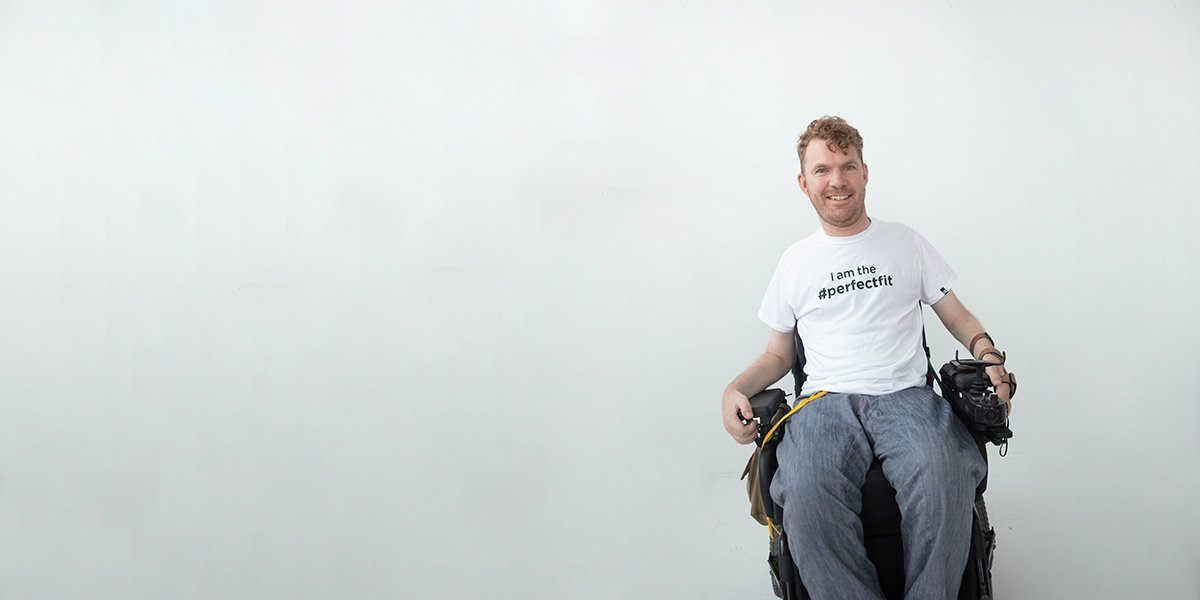 Full shot, seated. Luke is a model using a power chair. He wears gray jeans and a white t-shirt that reads, “I am the #PerfectFit.”
