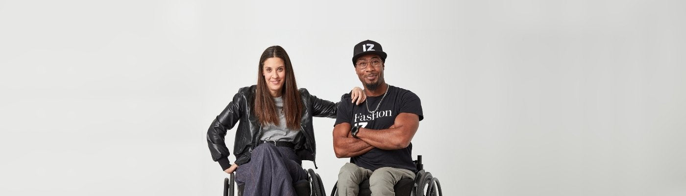 A woman using a manual wheelchair sits to the left of a man using a manual wheelchair, with her arm on his shoulder. She is wearing a leather jacket layered on top of a denim long skirt. He is wearing a black t-shirt layered on top of grey chinos.