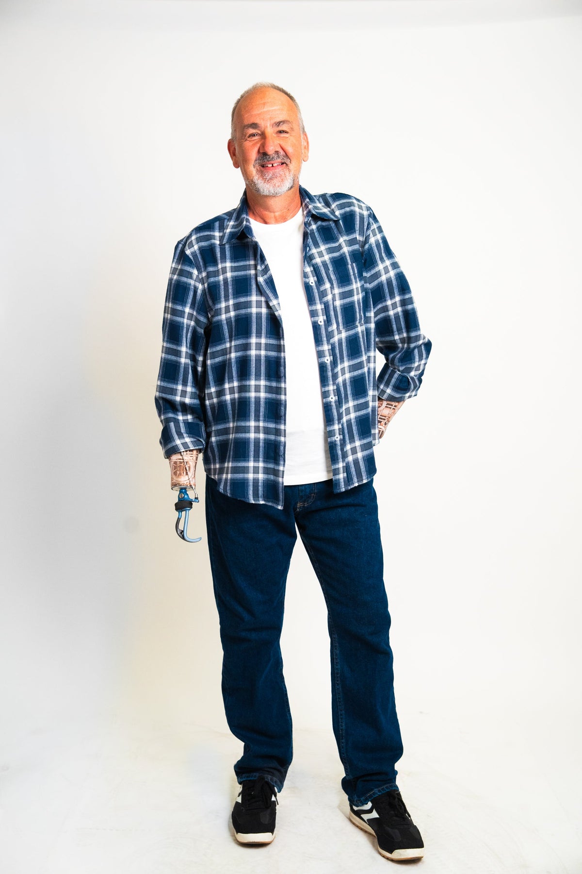 A man wearing a blue and white plaid flannel shirt over a white t-shirt, dark blue jeans, and black sneakers, standing against a plain white studio background.