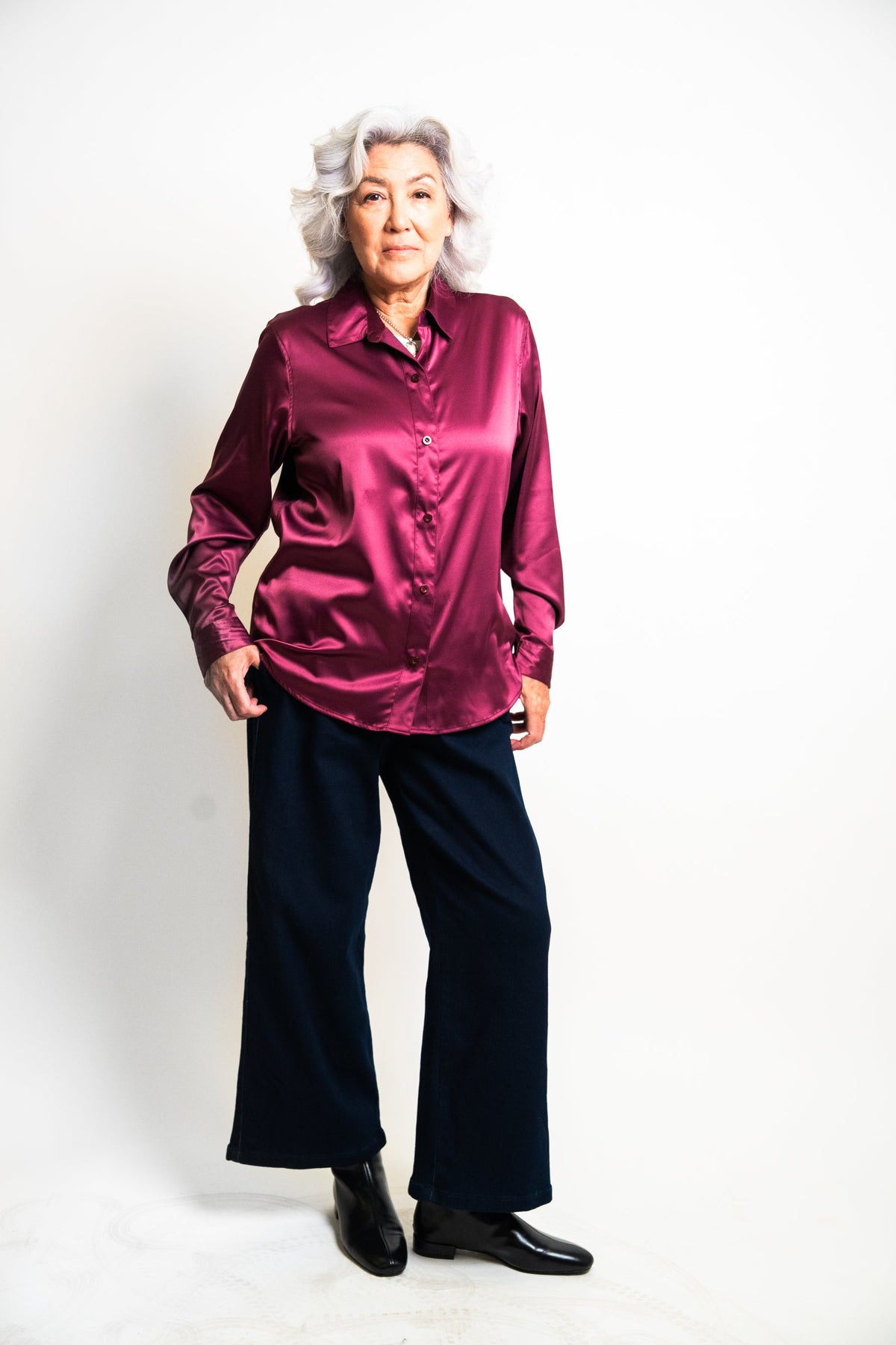 An older woman with gray hair stands against a plain white studio background wearing a shiny magenta-purple satin button-up shirt and dark navy wide-leg trousers with black shoes.