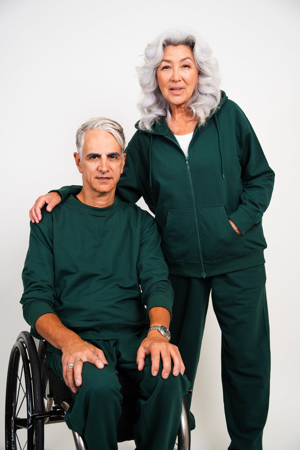 An elderly couple posing in matching dark green athletic tracksuits. The man is seated in a wheelchair, and the woman stands behind him with her arm around his shoulder.