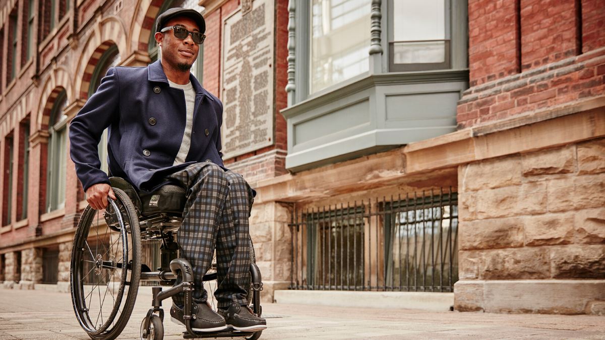 Man using manual wheelchair travels down city sidewalk with neighboring brownstone buildings. He wears an autumnal outfit including navy peacoat, checkered slacks and driver’s cap.