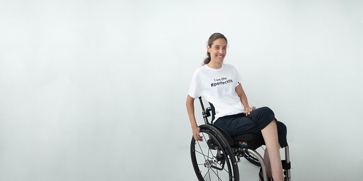 Partial side angle, seated. Anita is an individual using a manual wheelchair. She smiles as she crosses 1 leg over the other and rests 1 hand on her back wheel. Anita is wearing black capri pants and a white t-shirt that reads, “I am the #PerfectFit.”