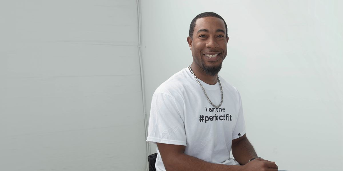 Medium shot, partial side angle, seated. Anthony smiles and rests both hands on his lap. He turns slightly towards camera, showing the text on his white t-shirt which reads “I am the #PerfectFit.”