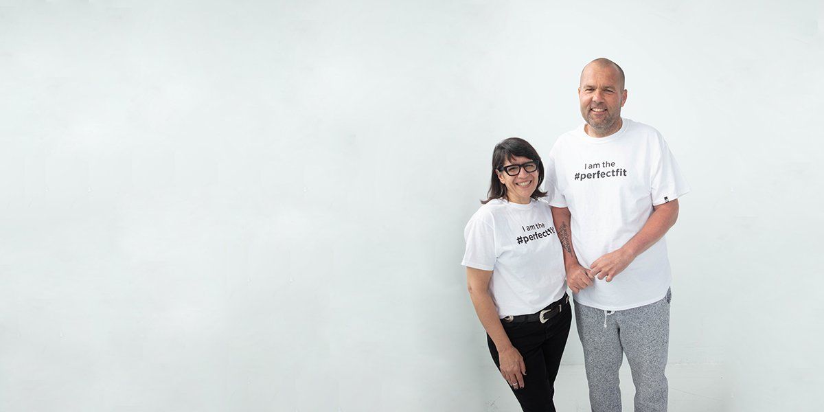 IZ Adaptive founder Izzy Camilleri standing with Chris. Both of them wear white t-shirts that reads, “I am the #PerfectFit.” Izzy wears black jeans with a black belt and Chris wears gray sweatpants with drawstring.