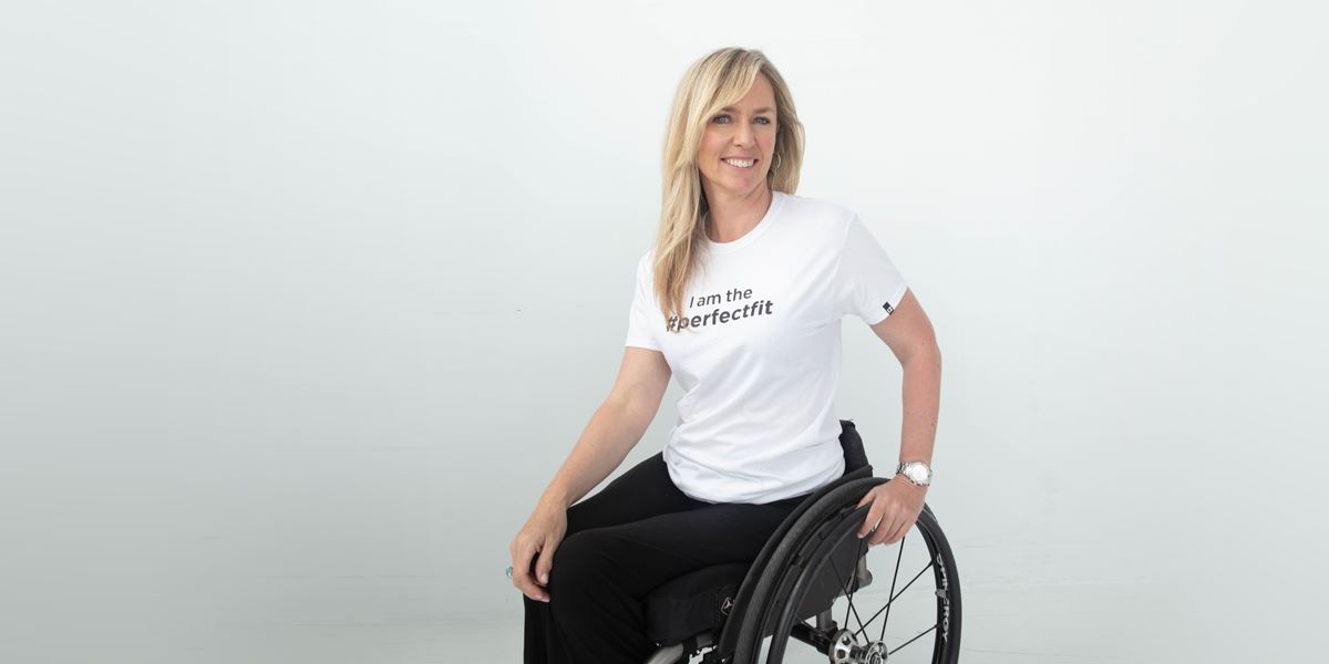 Partial side angle, seated. Joanne is an individual using a manual wheelchair. She rests 1 hand on the back wheel, twisting her torso towards camera. She wears black slacks and a white t-shirt that reads, “I am the #PerfectFit.”