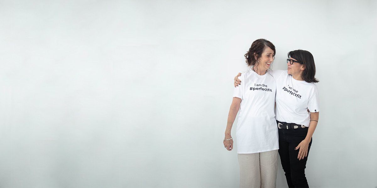 Medium shot, standing. Jodi and IZ Founder Izzy Camilleri smile as they wrap an arm around each other. Both of them wear a white t-shirt that reads, “I am the #PerfectFit.” Jodi wears beige jeans and Izzy wears black jeans with a black belt.