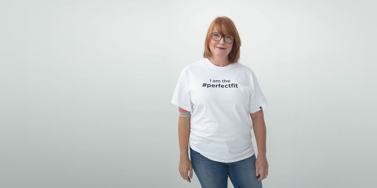 Medium shot, standing. Mary Anne is an individual using a transhumeral prosthesis. She wears light blue denim jeans and a white t-shirt that reads, “I am the #PerfectFit.”