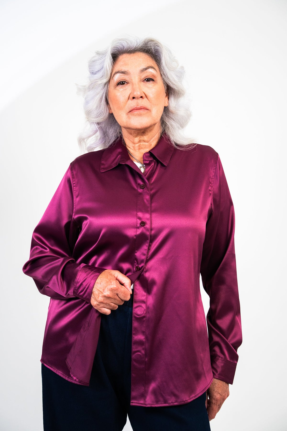 An older woman with gray hair wearing a shiny magenta-purple satin button-up shirt and dark pants, standing against a plain white studio background.