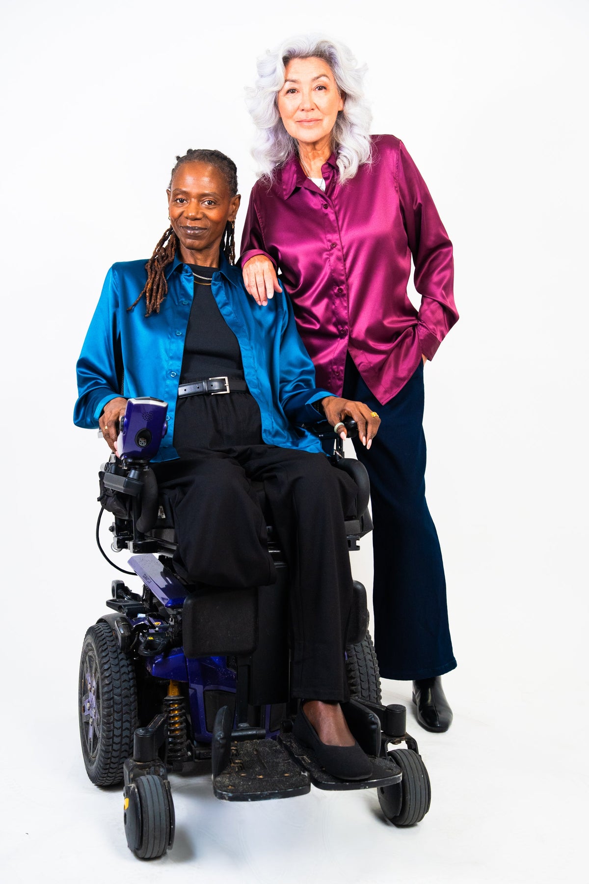 A woman in a blue satin jacket sitting in a motorized wheelchair and a woman in a shiny magenta satin blouse standing beside her, photographed against a plain white studio background.