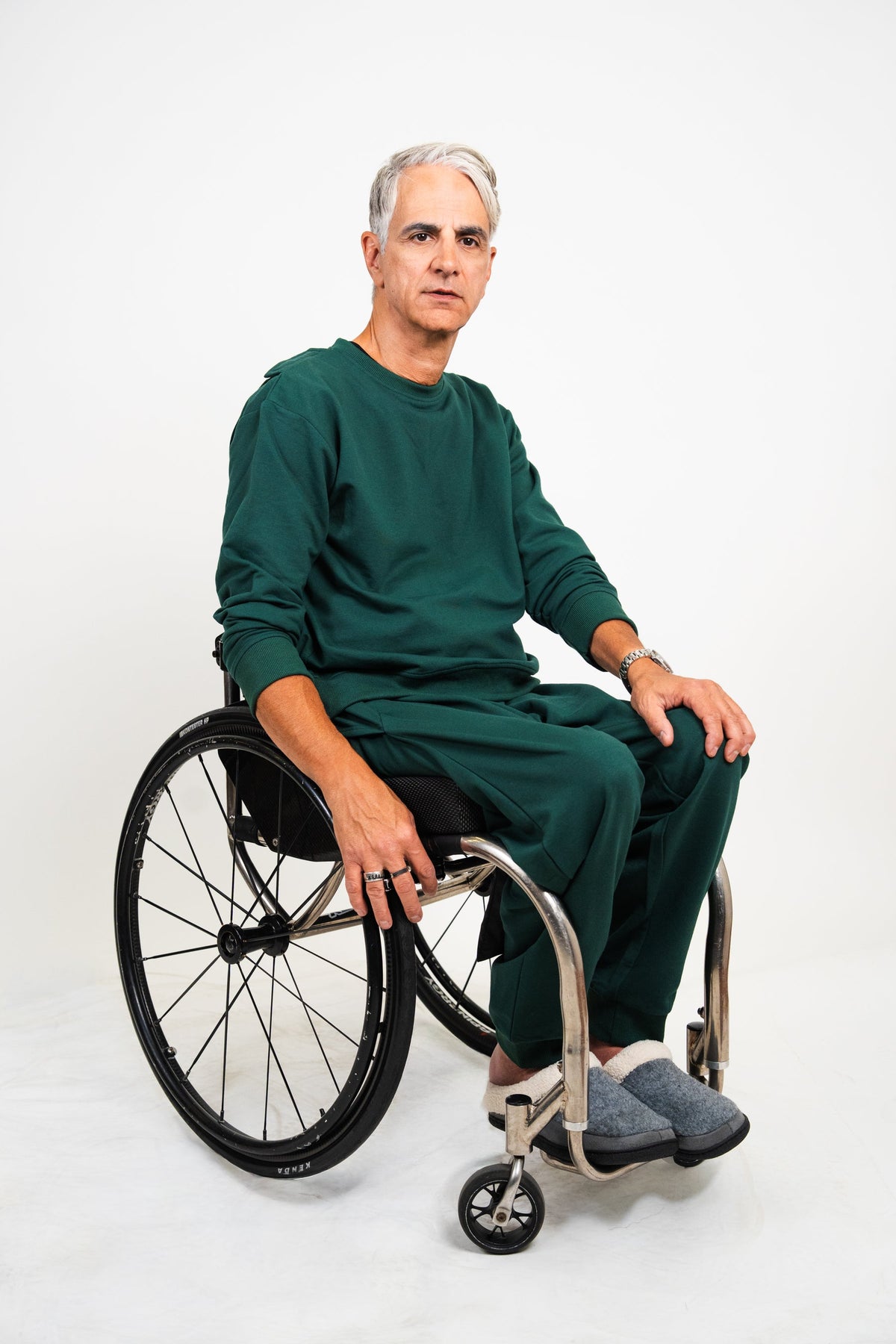 A man with gray hair sits in a wheelchair wearing a matching dark green long-sleeve top and pants set, with gray slip-on shoes, against a plain white studio background.