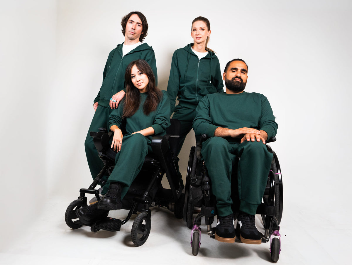 Four people wearing matching dark green athletic tracksuits posed together in wheelchairs against a plain light gray studio background.