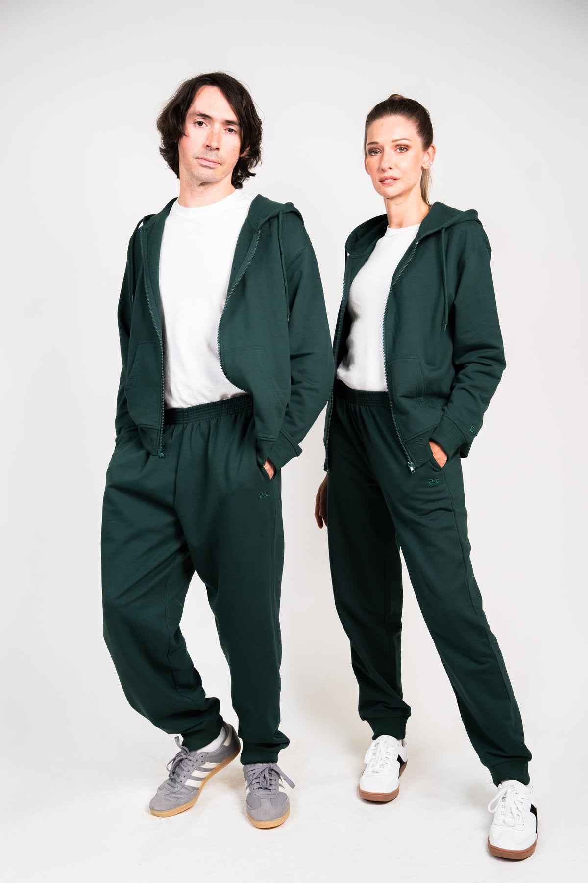 A man and a woman modeling matching dark green hooded sweatshirt and jogger pants sets, styled over white t-shirts and white sneakers, against a plain light gray studio background.